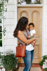 A mother holds her baby and wears a stylish leather tote bag outside her home.
