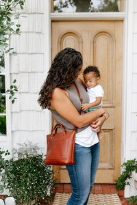 A mother holds her baby and wears a stylish leather tote bag outside her home.
