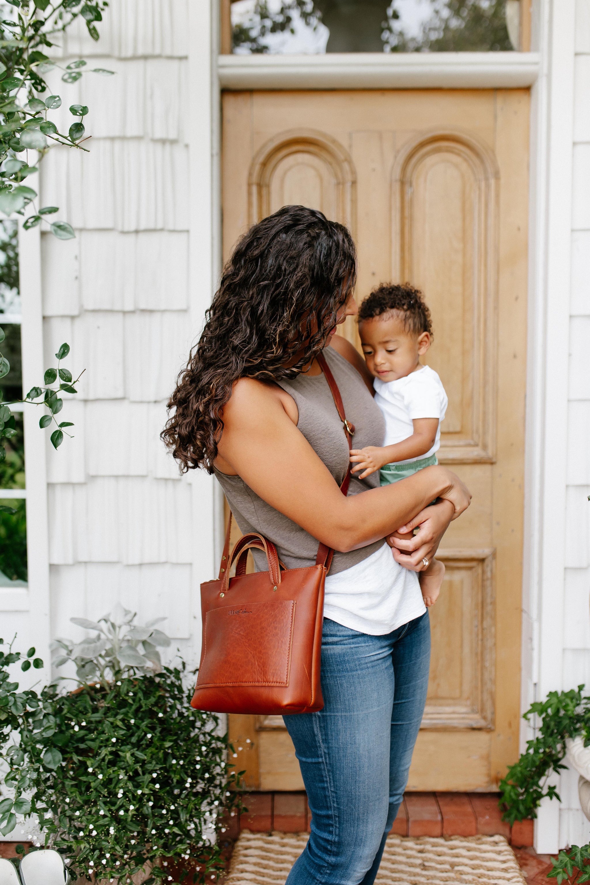 A mother holds her baby and wears a stylish leather tote bag outside her home.

