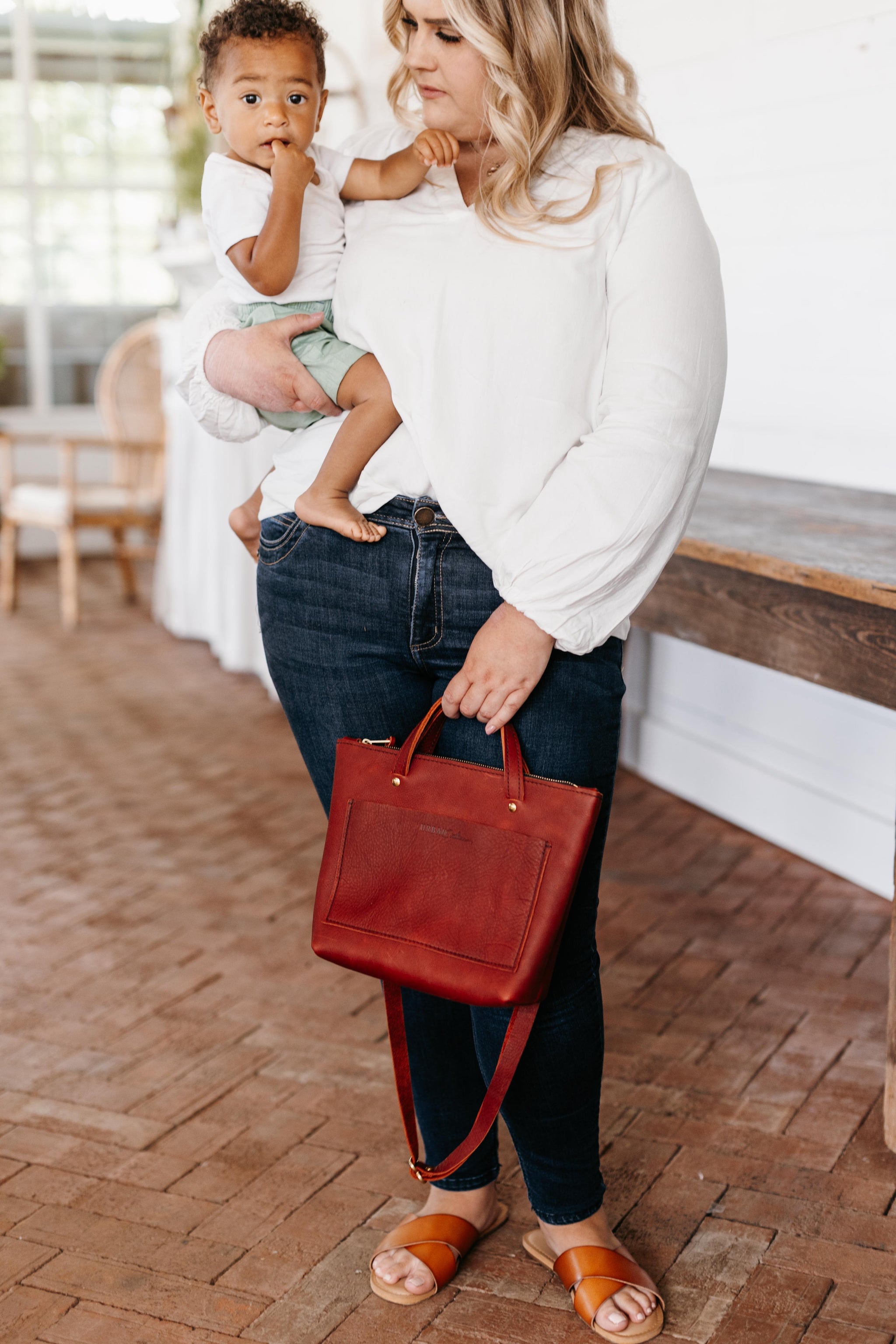 A woman carries a cute toddler while holding a stylish red leather tote bag.

