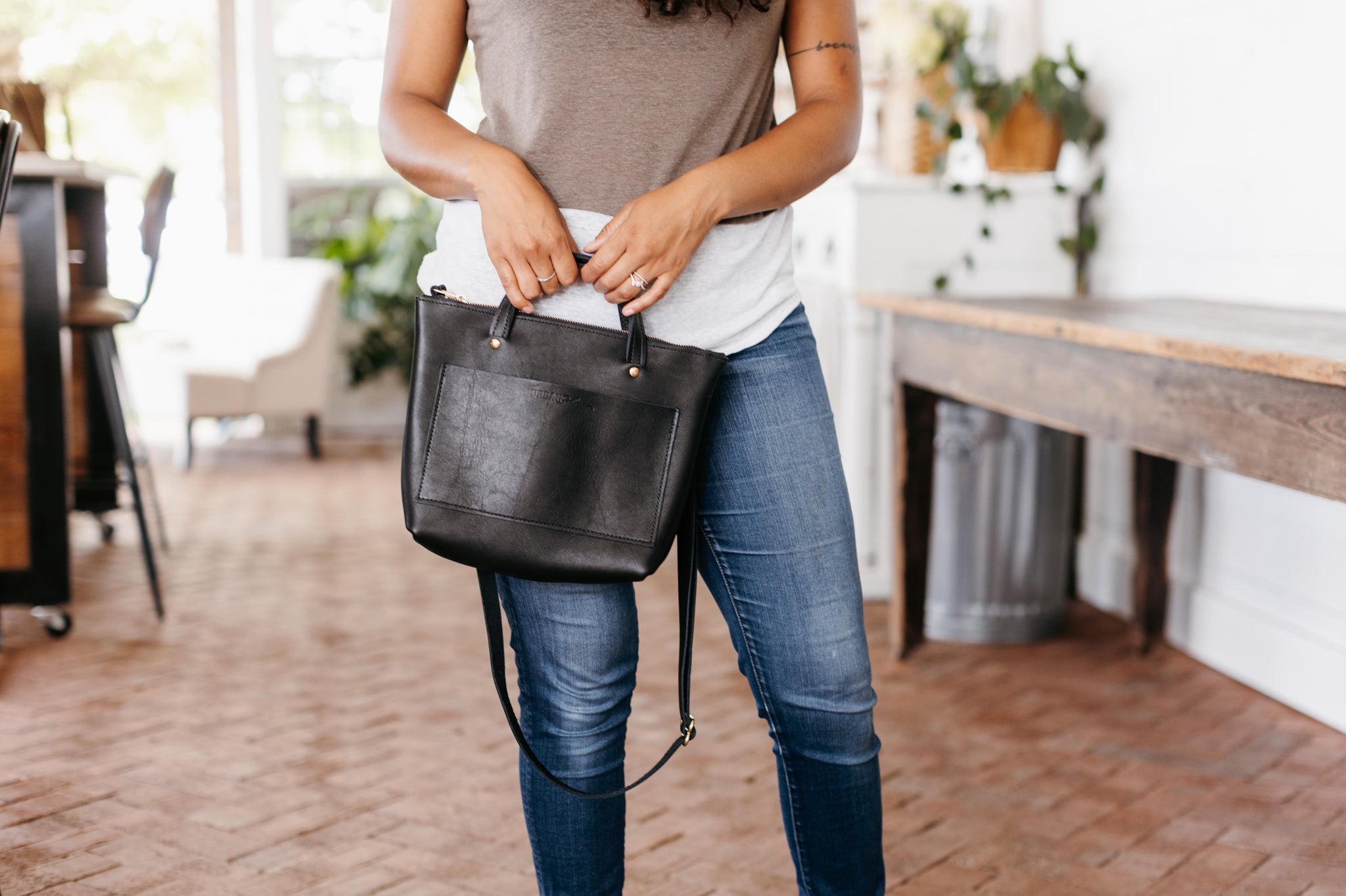 Woman holds a stylish black leather tote bag indoors, showcasing its classic design and versatility.
