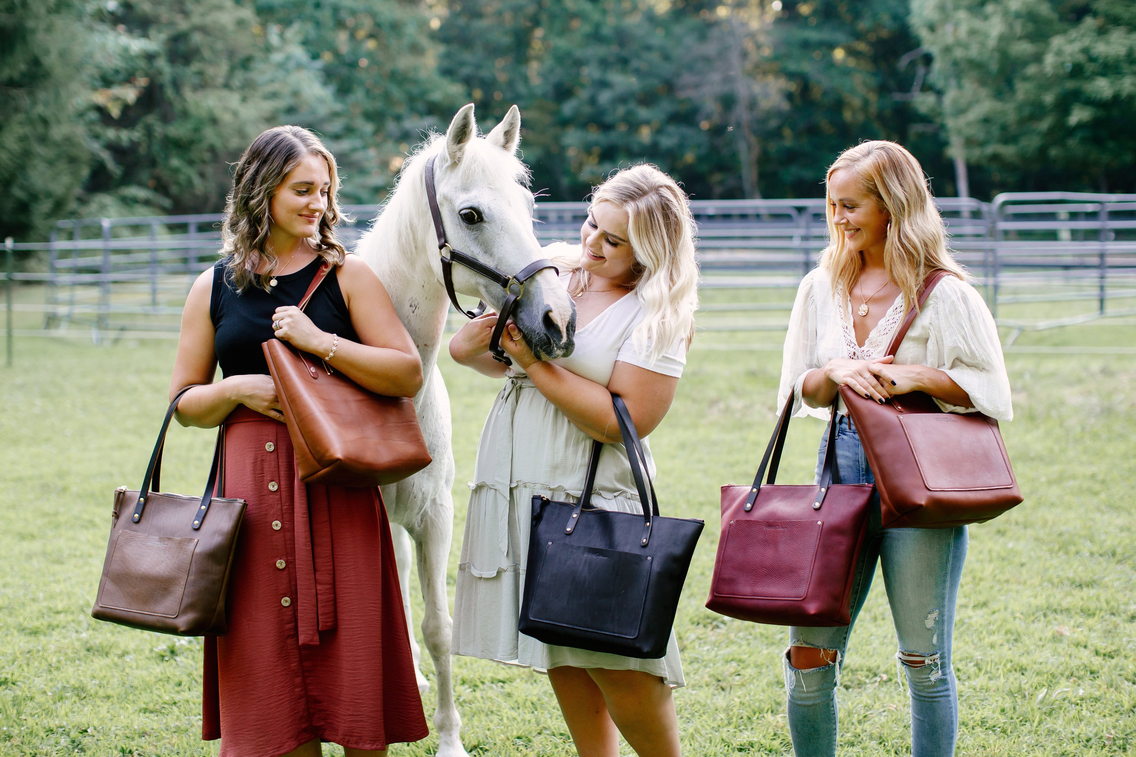 Three stylish women pose with a white horse, showcasing their handcrafted leather tote bags in a grassy field.
