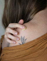 A woman applies natural healing balm to her shoulder with a delicate botanical tattoo.
