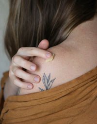 A woman applies natural healing balm to her shoulder with a delicate botanical tattoo.

