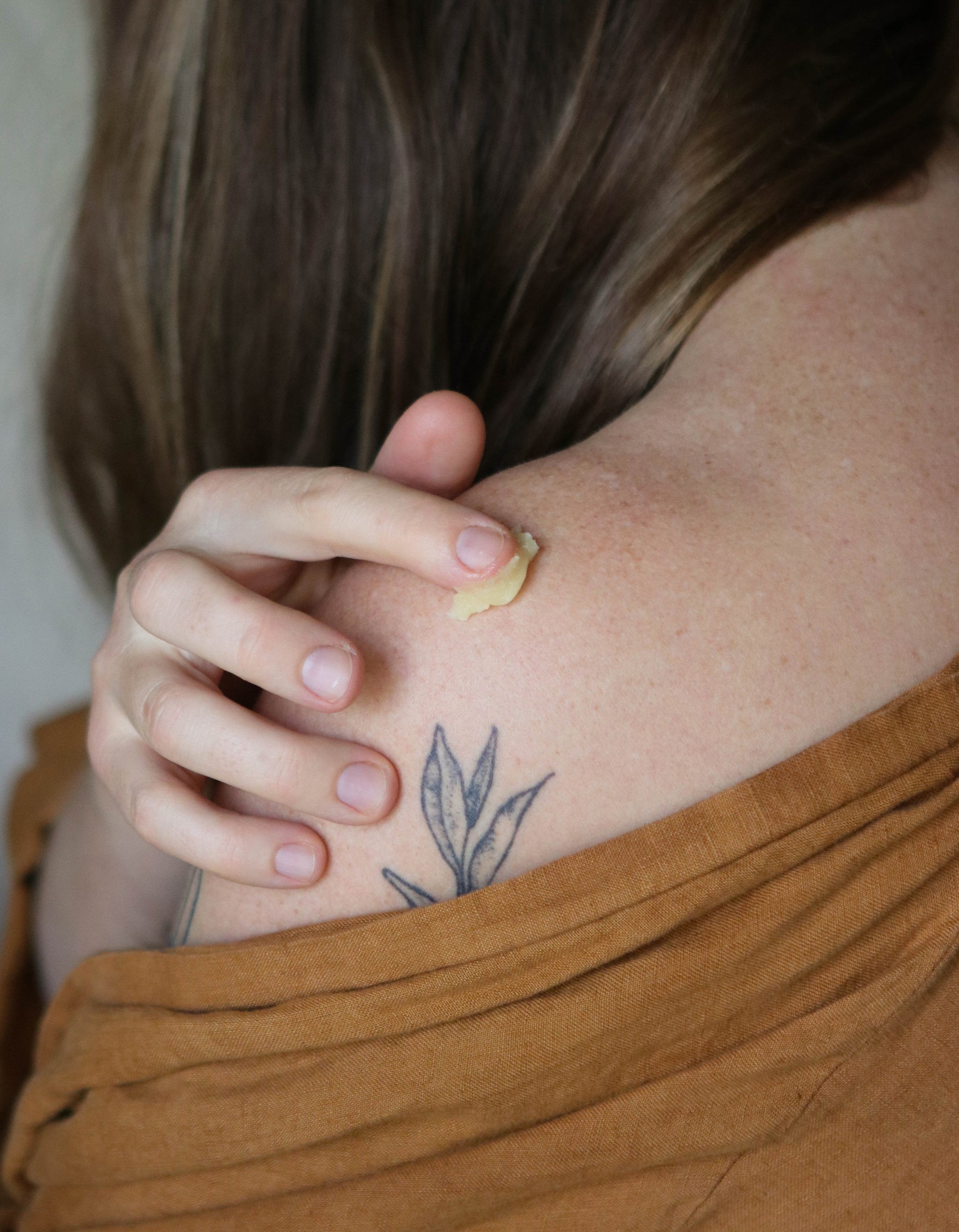 A woman applies natural healing balm to her shoulder with a delicate botanical tattoo.
