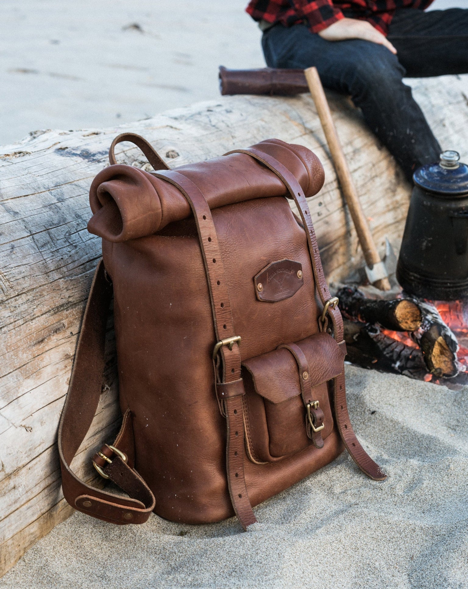 Brown leather Urban Southern backpack on a log with a person sitting by a fire in the background