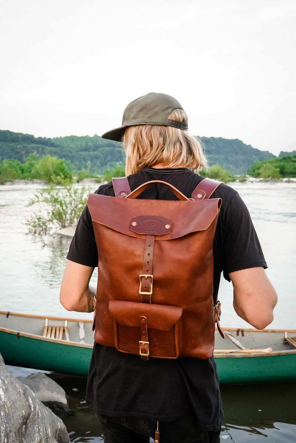 Man with a brown leather Urban Southern backpack standing by a body of water with a canoe.