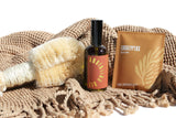 Showcases a dry brush, anointing oil, and eucalyptus bath soak from URB.APOTHECARY on a textured, fringed blanket.
