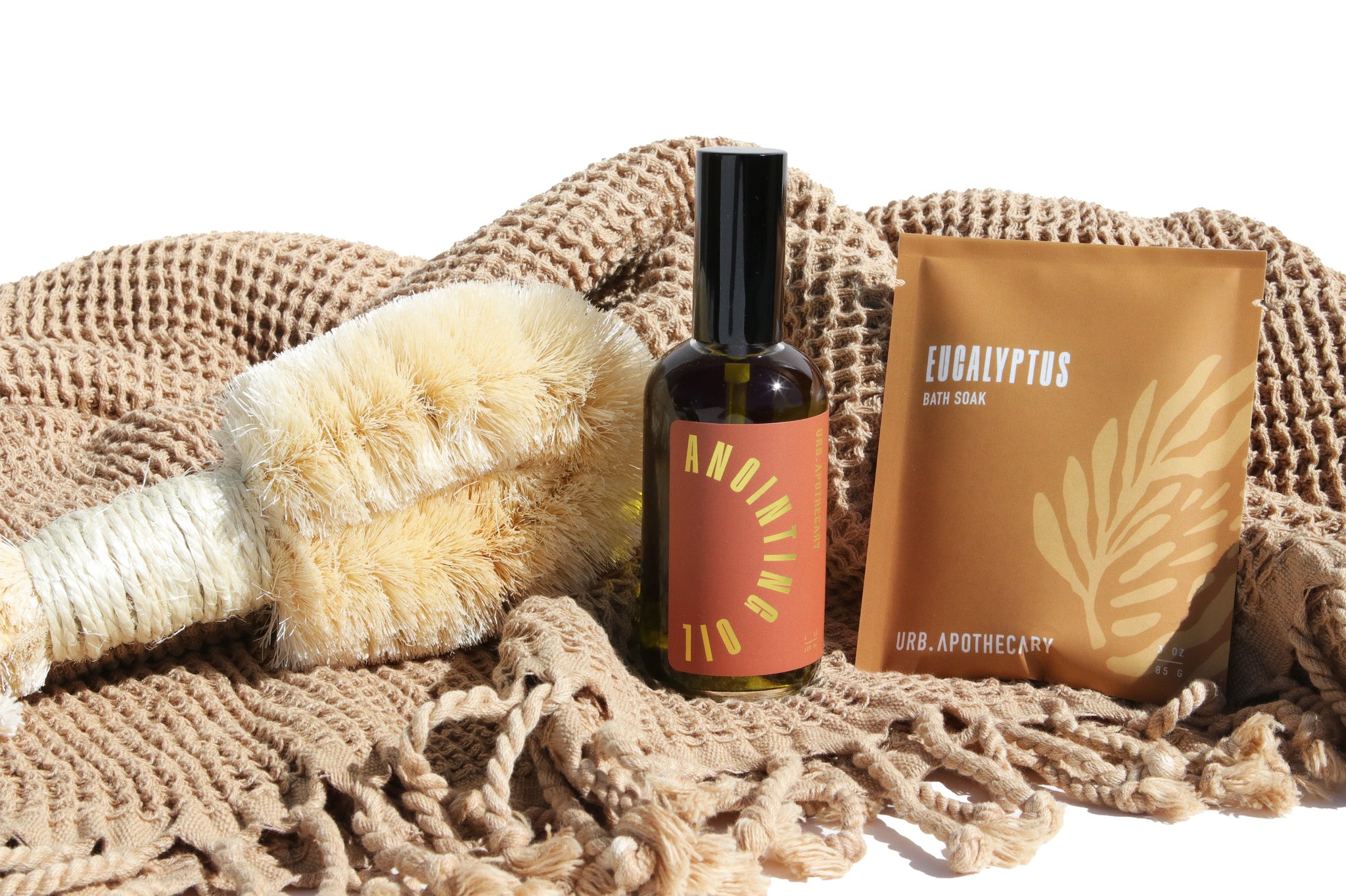 Showcases a dry brush, anointing oil, and eucalyptus bath soak from URB.APOTHECARY on a textured, fringed blanket.
