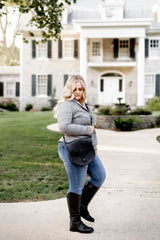 Woman in jeans and riding boots carries a saddle bag in front of a large house.
