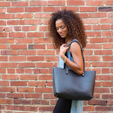 A woman with curly hair carries a stylish black leather tote bag against a brick wall.
