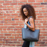 A woman with curly hair carries a stylish black leather tote bag against a brick wall.
