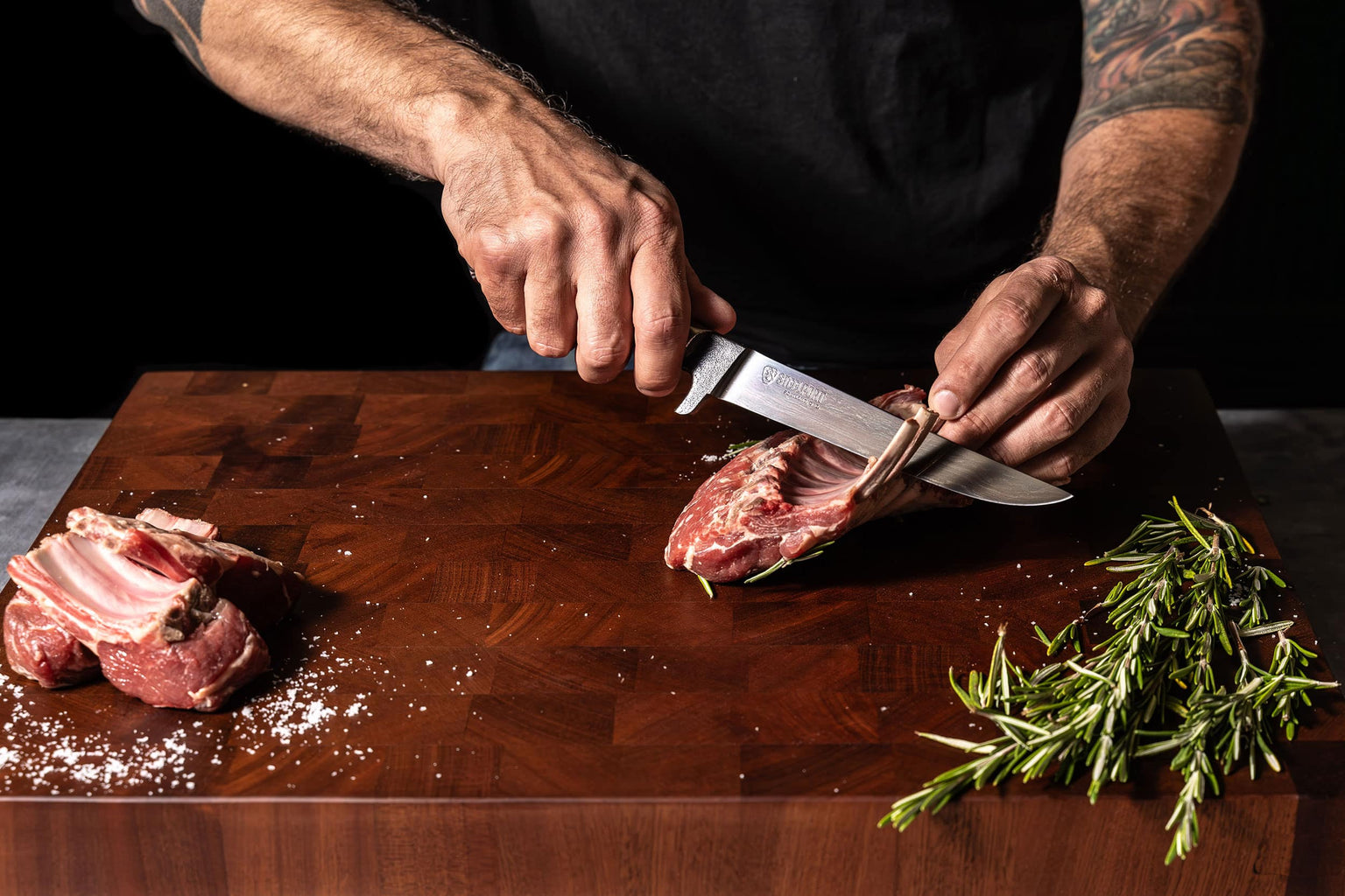 Person cutting raw meat on a wooden cutting board with a Steelport knife, surrounded by rosemary.