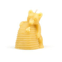 Close-up shows a golden beeswax bear candle hugging a beehive on a white background.
