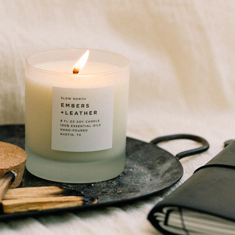 Candle labeled 'Embers + Leather' on a dark surface with a soft background