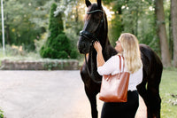 A blonde woman adjusts the bridle of a beautiful black Friesian horse during an equestrian photo shoot.
