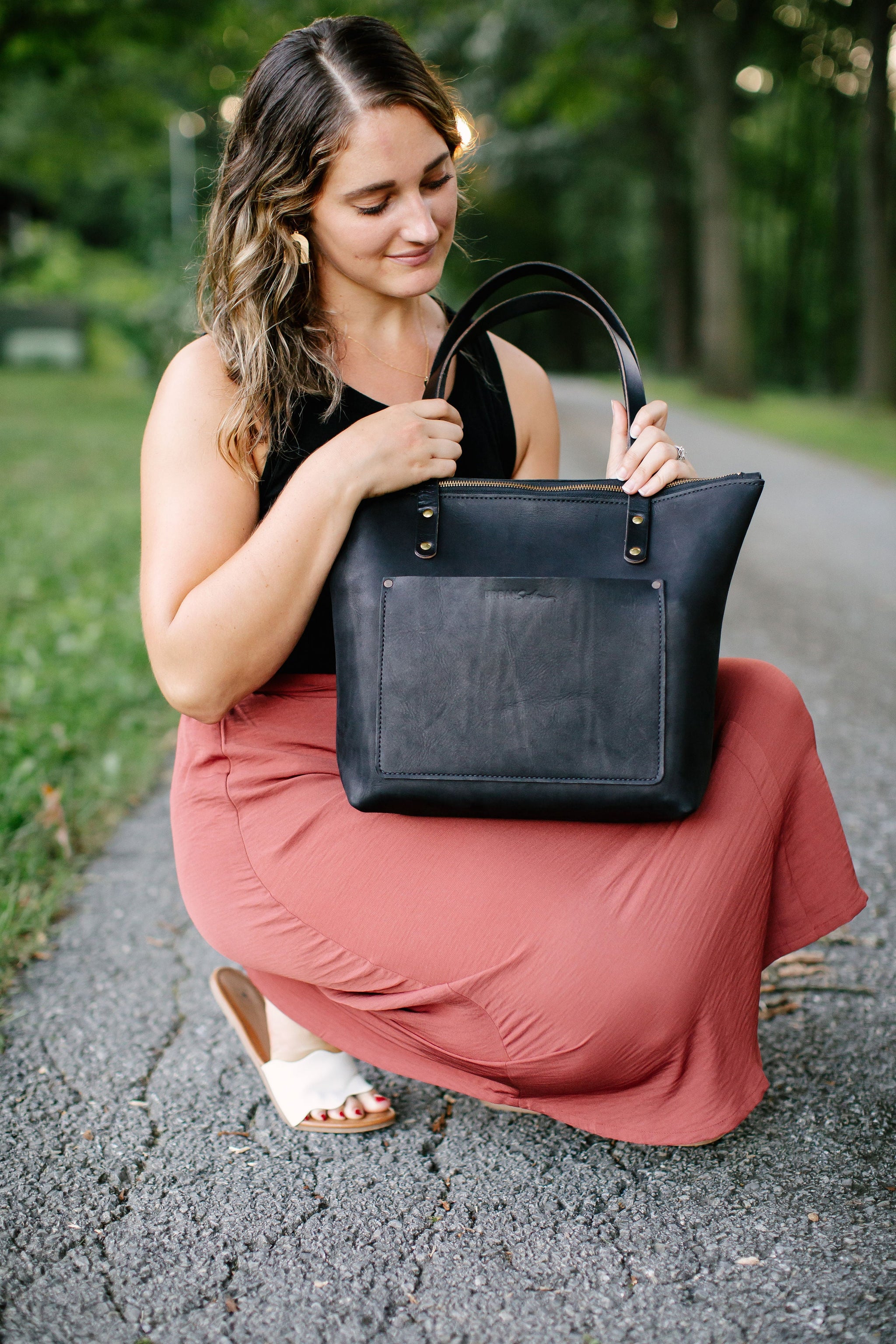 A woman kneels on a path and admires her new leather tote bag.
