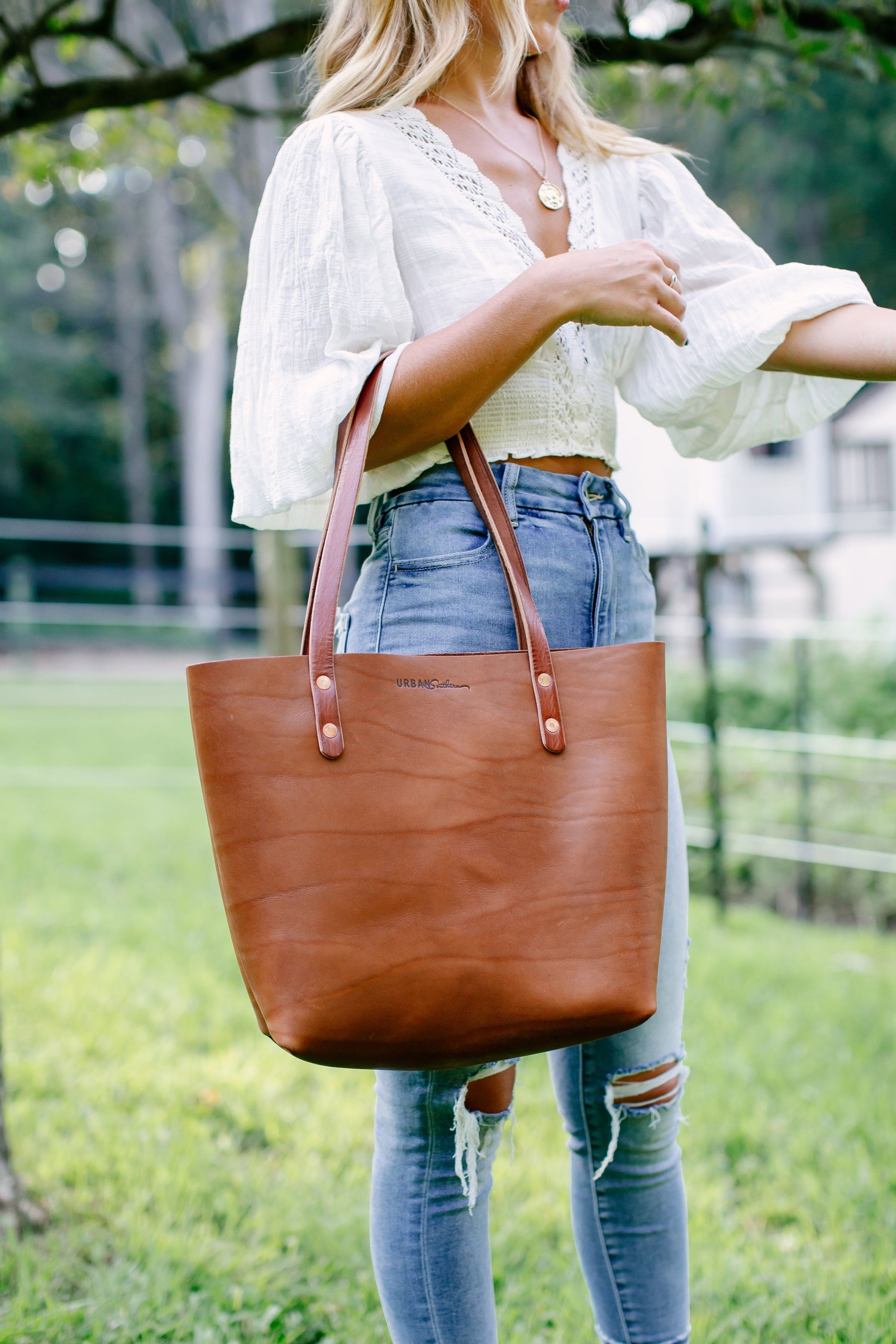 A woman showcases her stylish outfit and carries a brown leather tote bag in a grassy outdoor setting.
