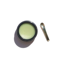 A jar of green healing balm sits beside a small golden spatula applicator.
