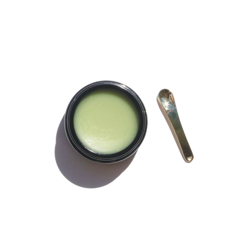 A jar of green healing balm sits beside a small golden spatula applicator.
