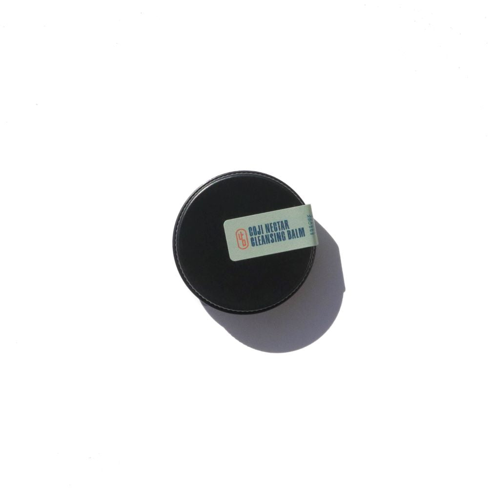 A black jar of Goji Nectar cleansing balm sits on a white surface.
