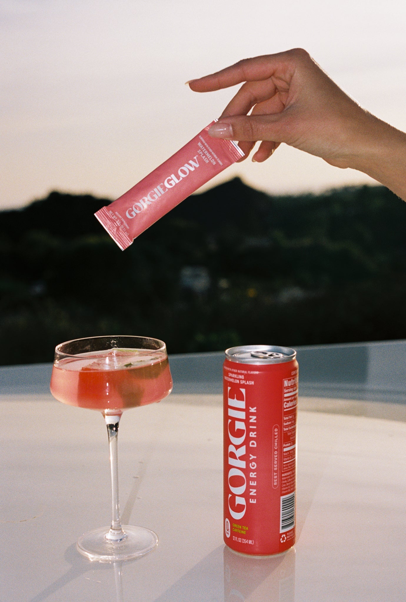 Hand holding a Gorgie drink package above a glass with a sunset background
