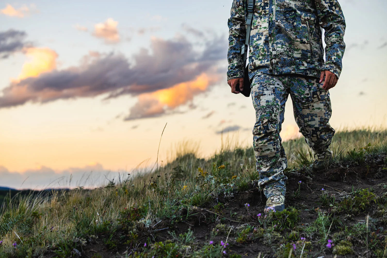 Person in camouflage clothing standing on a grassy hill with a sunset sky.
