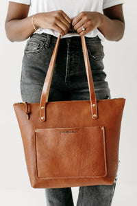 A person showcases a stylish, handcrafted brown leather Urban Southern tote bag with its leather straps and front pocket detail.
