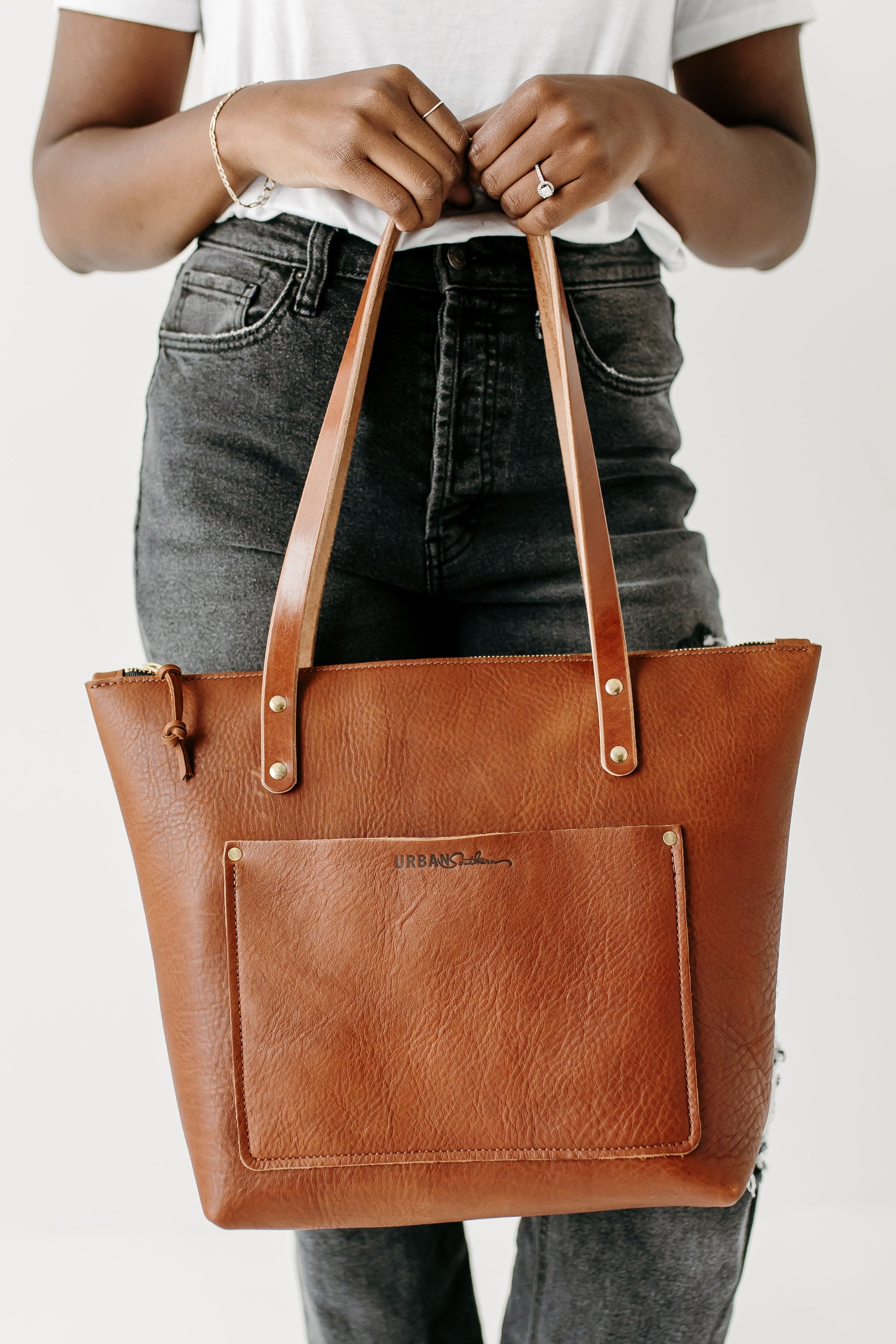 A person showcases a stylish, handcrafted brown leather Urban Southern tote bag with its leather straps and front pocket detail.
