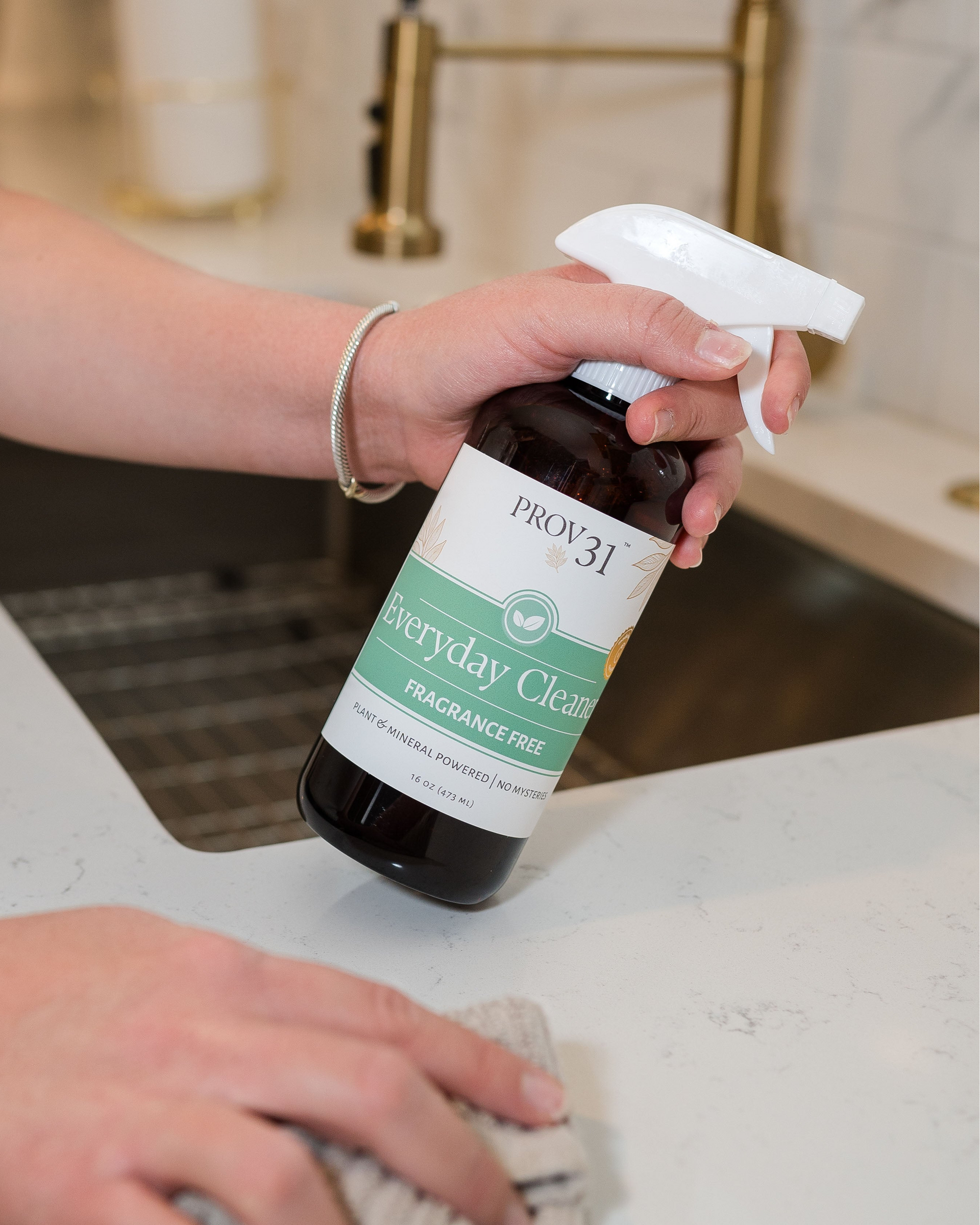 A person holds a bottle of PROV31 Everyday Clean fragrance-free spray over a white marble kitchen counter, with a sink and faucet blurred in the background.