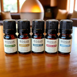 Rogue Evergreens presents a collection of five small-batch essential oils, including Douglas Fir, Ponderosa Pine, Grand Fir, Incense Cedar, and Noble Fir, displayed on a wooden surface.
