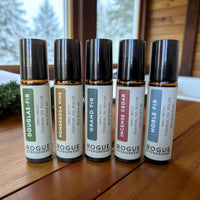 Rogue Evergreens presents a collection of small-batch essential oil roller bottles, including Douglas Fir, Ponderosa Pine, Grand Fir, Incense Cedar, and Noble Fir, arranged on a wooden surface with a snowy window in the background.
