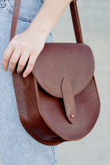 A woman wearing jeans carries a stylish brown leather saddlebag with a brass closure detail.
