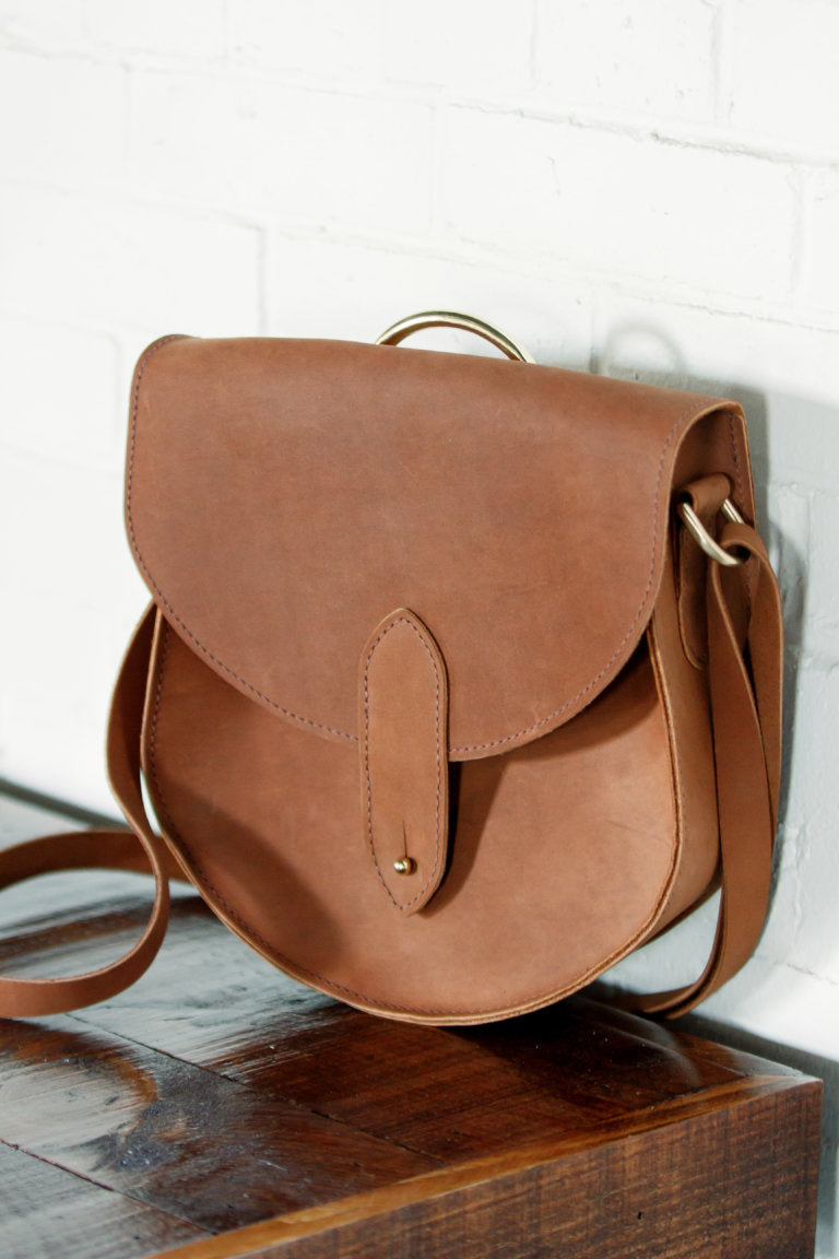 A brown leather saddlebag purse with a gold handle sits on a dark wooden surface against a white brick wall.
