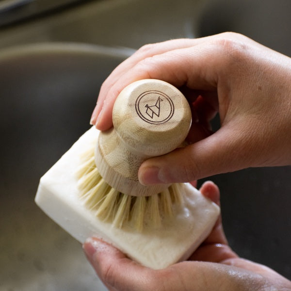Hands scrub a white dish soap bar with a bamboo dish brush over a sink.
