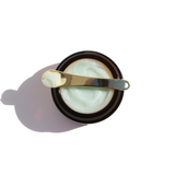 A jar of light green face cream sits with a gold applicator spoon on top.
