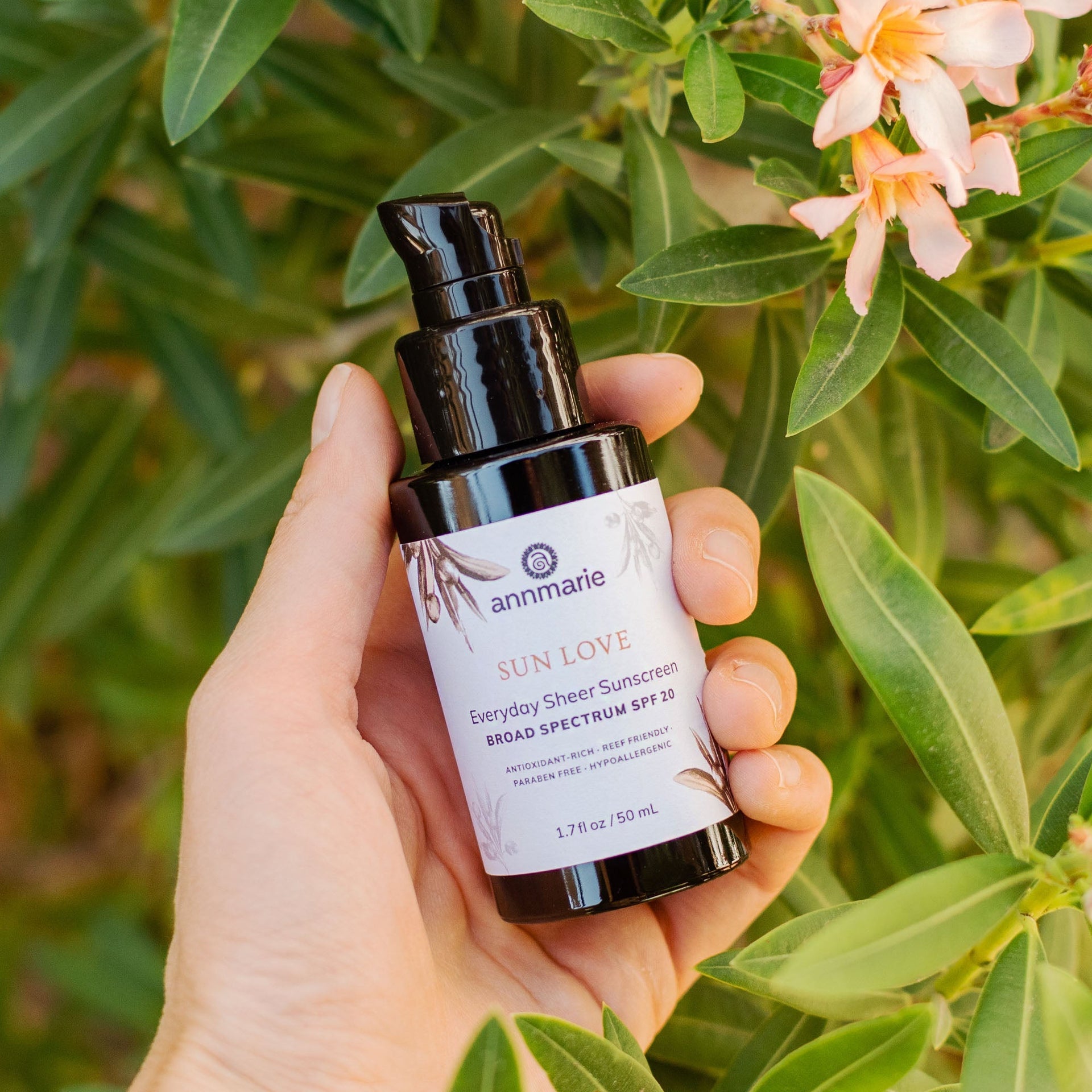 A hand holds Annmarie Sun Love sheer sunscreen SPF 20 in front of a leafy green plant with peach-colored flowers.
