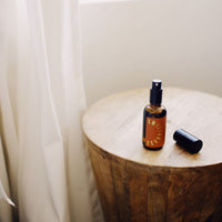 Still life shows a Sweetgrass aromatherapy spray bottle resting on a wooden side table beside a white curtain.
