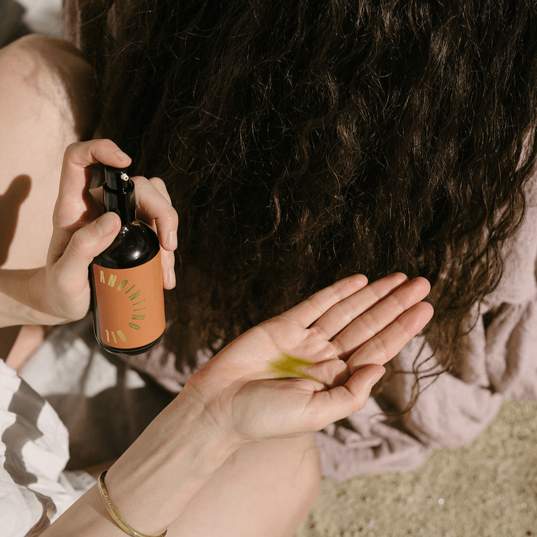 A woman sprays "Anointing Oil" hair product onto her hand before applying it to her curly hair.
