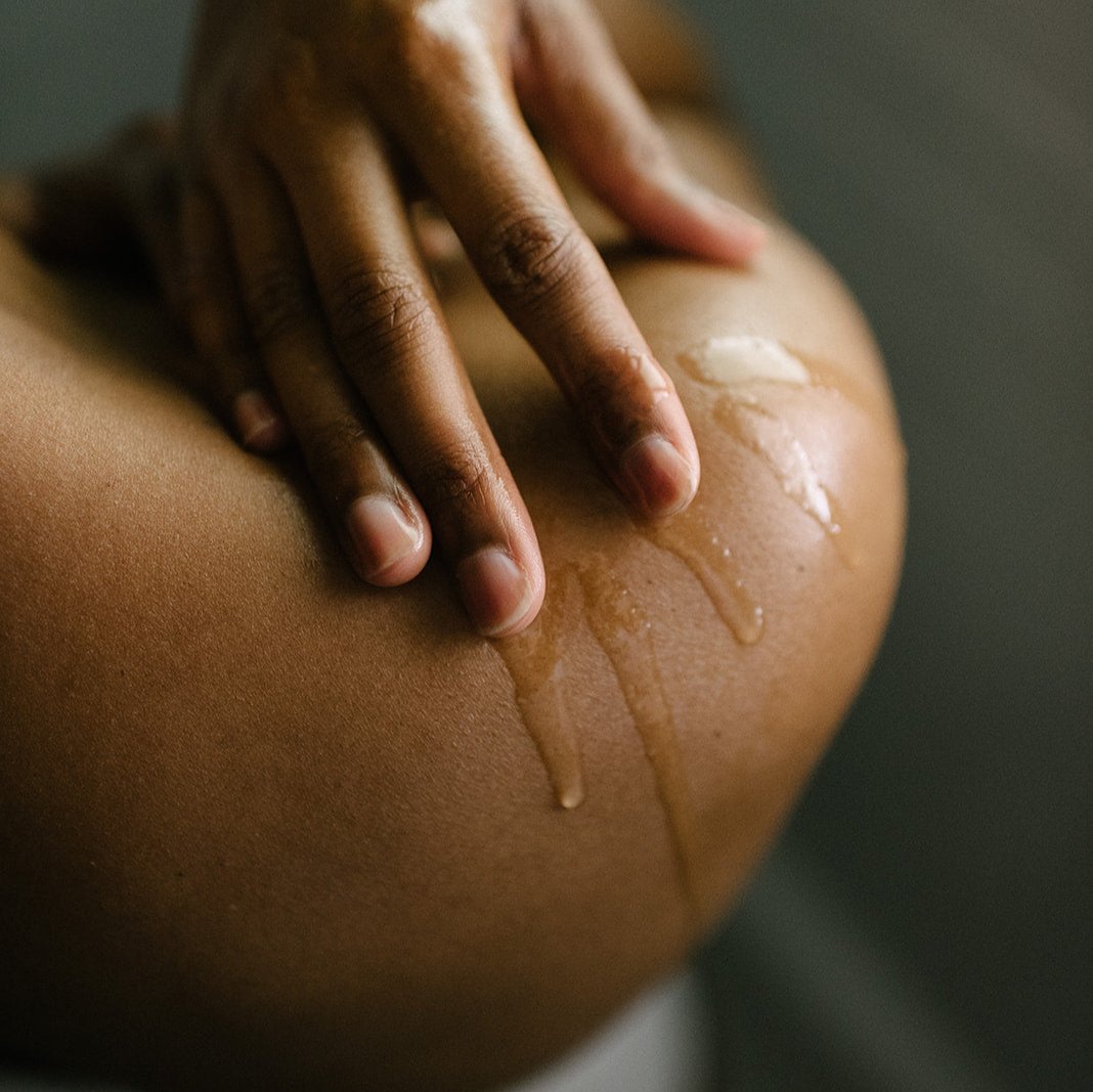 A hand applies moisturizing body oil to tanned skin, leaving a dewy glow.
